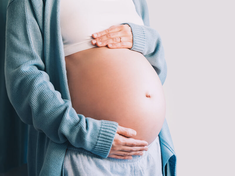 Schwangere Person hält mit beiden Händen ihren runden Bauch in heller Kleidung